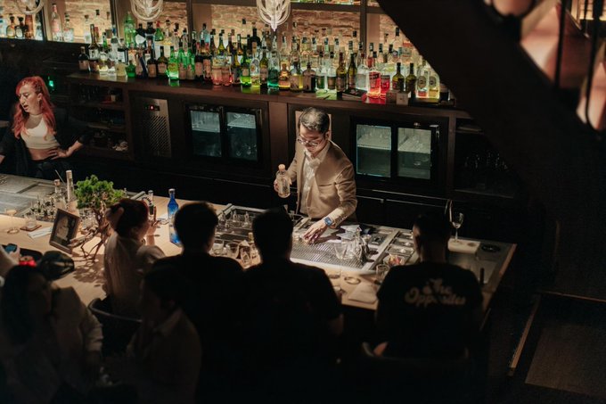 A bartender in a tan jacket pours a cocktail behind a well-stocked bar while patrons watch, with one wearing a "York Apparel" shirt. The post's playful reference to "whipping up a buzz" for smooth Bitcoin charting cleverly pairs with the image of mixology, suggesting that keeping BTC's momentum flowing requires a careful, skilled hand.
