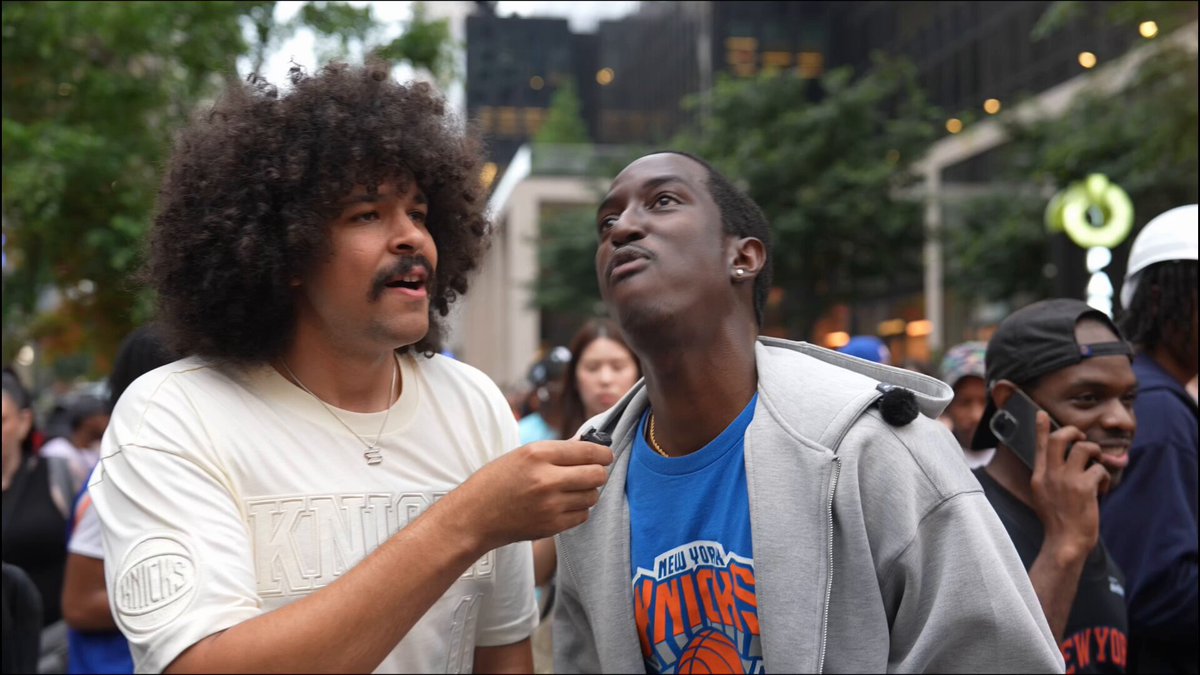 The image shows two men standing next to each other, with one man pointing at something above his head while the other looks up. The background includes a crowd of people gathered together, suggesting that they might be attending an event or watching a game related to the New York Knicks basketball team. There is also a post on social media about the current state of the stock market and the word "Solana" appears in the image.