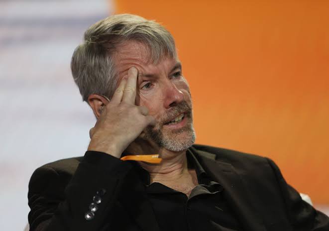 A man with gray hair and a beard, wearing a black jacket, gestures near his temple while speaking at what appears to be a conference or interview setting with an orange background. The image relates to the post as it likely depicts Michael Saylor, executive chairman of MicroStrategy, discussing the company's ongoing Bitcoin accumulation strategy.