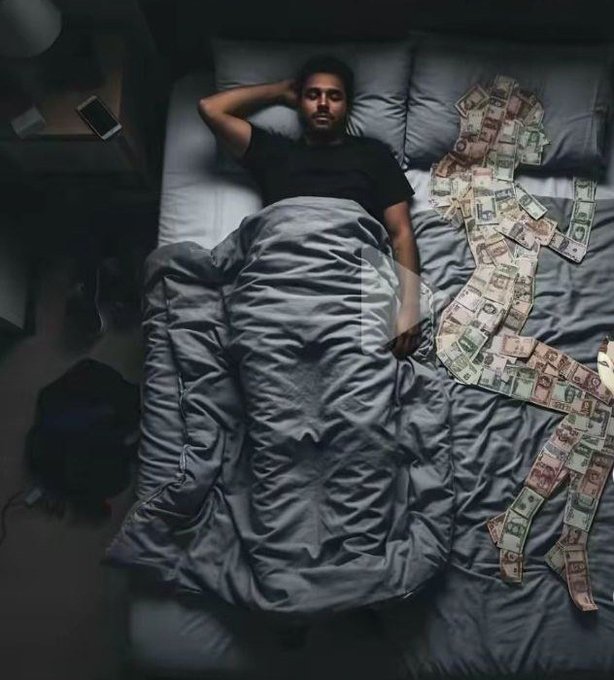 A man lies in bed under a gray duvet with a large pile of euro and dollar banknotes scattered across the mattress beside him.