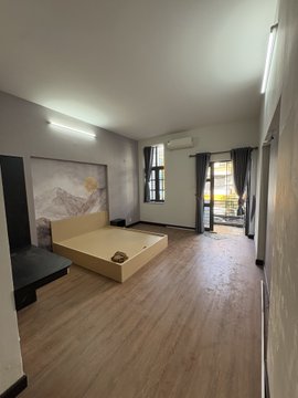 An empty bedroom with light wood flooring, featuring a wooden bed frame, a wall-mounted mountain landscape artwork, an air conditioner, and a balcony with dark curtains.