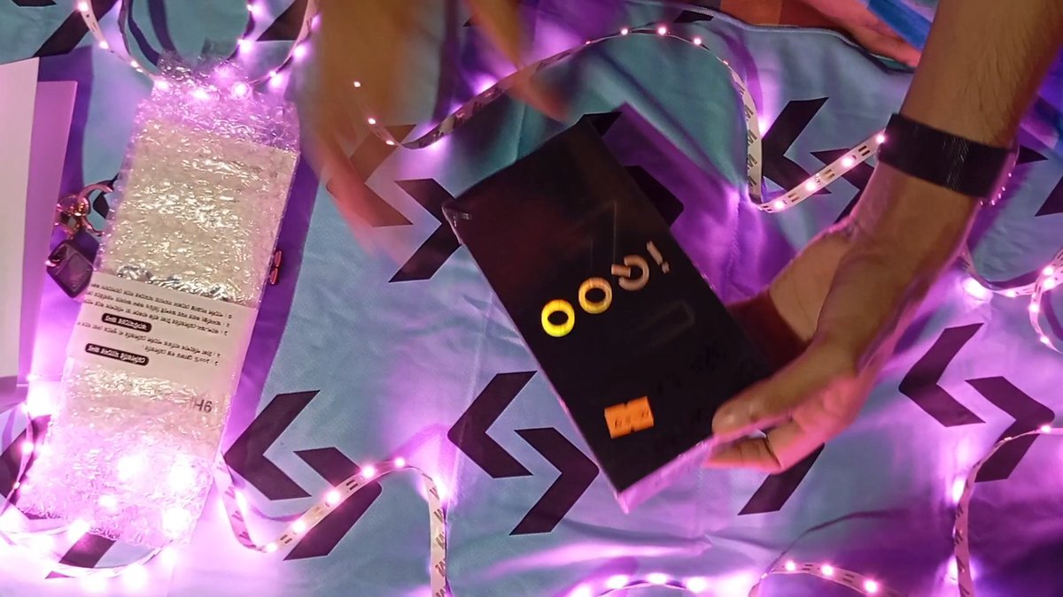 A pair of hands is unboxing a Poco phone on a patterned bedsheet surrounded by purple LED string lights, with a bubble-wrapped package visible nearby.