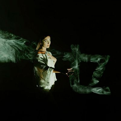 A dark, atmospheric double-exposure image of a woman in white interacting with swirling, ghostly green smoke or energy against a black background.