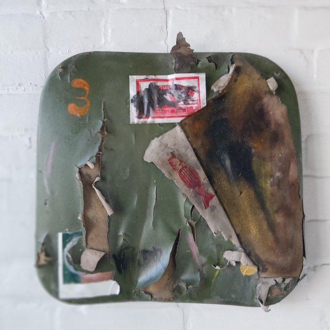 A weathered, damaged green metal sign mounted on a white brick wall, featuring torn and faded stickers, including one with the word 'TURK' and a red fish design.