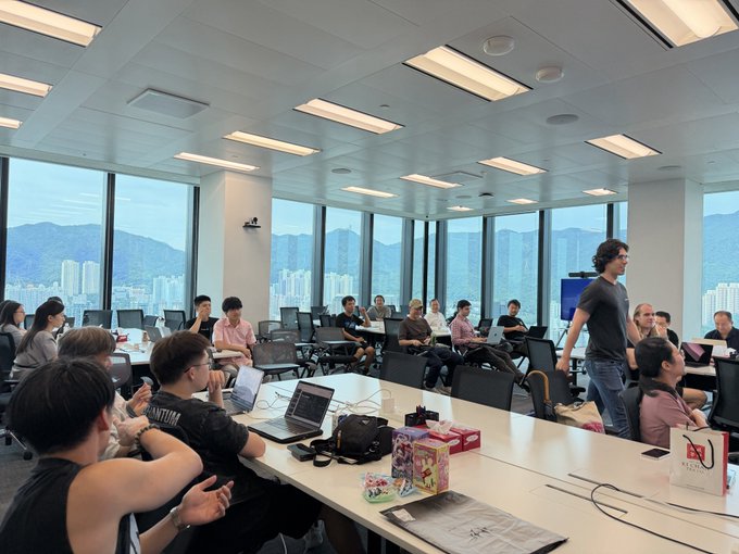 A group of people gather in a modern, window-lined office with laptops open, while one person stands at the front leading what appears to be a meeting or presentation.
