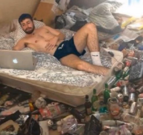 A shirtless man in dark underwear lies on a mattress on the floor surrounded by a massive amount of trash, empty bottles, and containers, with a silver laptop in front of him.