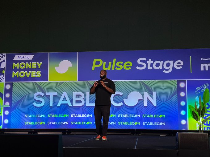 A man stands on a stage in front of a large screen displaying 'STABLECON', 'Pulse Stage', and 'Making MONEY MOVES', gesturing with his hands as if giving a presentation.