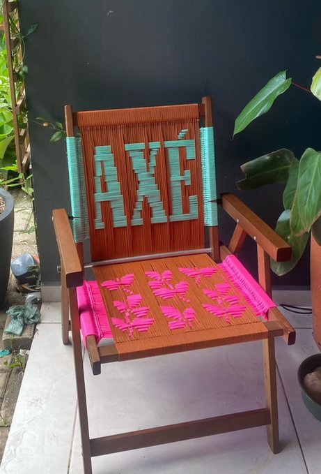 A wooden folding chair features handwoven thread designs in teal on the backrest and pink on the seat, displaying the letters 'HXC' against a backdrop of plants and a dark wall.