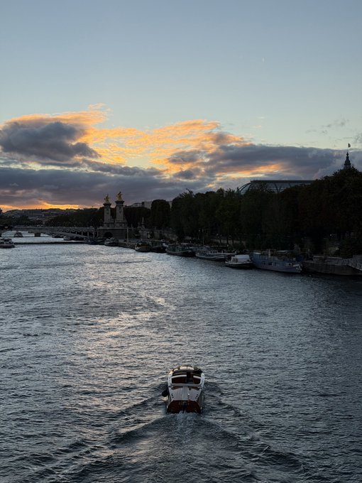 A scenic sunset view over the Seine River in Paris, showing a small boat navigating the water with the Pont Alexandre III bridge and its golden statues visible in the background.