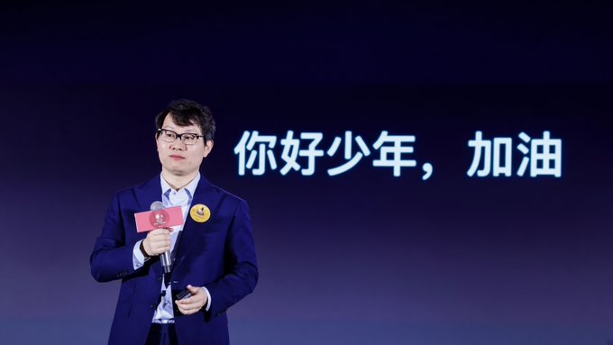 A man in a blue suit and glasses speaking into a microphone on stage, with Chinese text '你好少年，加油' (Hello youngster, keep going) displayed on the screen behind him.