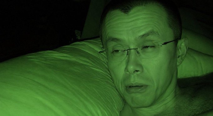 A close-up selfie of a man with glasses lying on a pillow, tinted green from night vision.