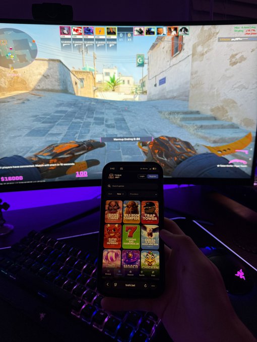 A person holds a smartphone displaying the "Stake" casino app with various slot games like "Joker Bombs," "Squid Game," and "Chicken" while playing Counter-Strike (CS:GO) on a large monitor in the background, with their orange-gloved character holding a karambit knife mid-round—reflecting the post's intent to gamble on slots during a gaming session due to losses on memecoins like Toshi.