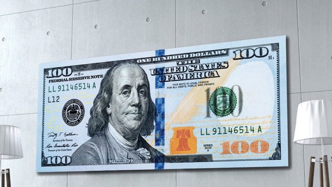A large canvas or print of a US $100 bill is mounted on a gray wall, flanked by white lamps on either side.