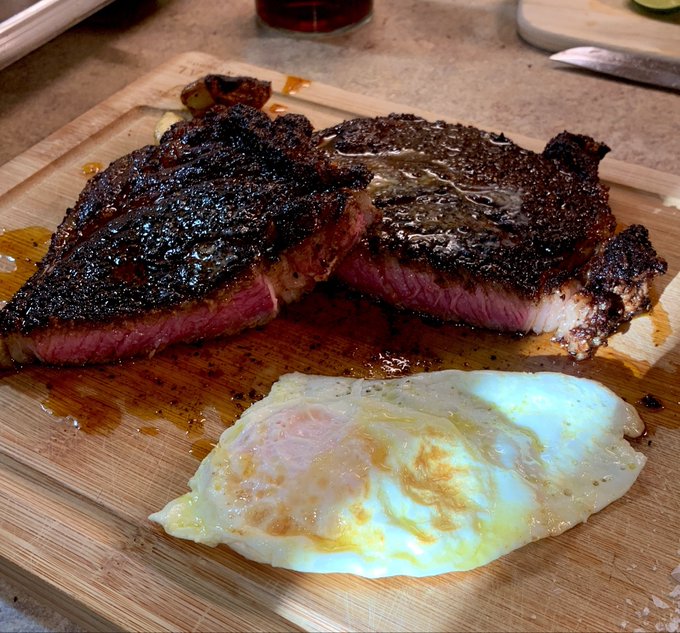 A perfectly seared T-bone steak with a rich, charred crust and pink medium-rare interior is served on a wooden cutting board alongside a sunny-side-up egg, with juices pooling around the meat—visually playing on the "halving" joke by showing the steak split into two halves.