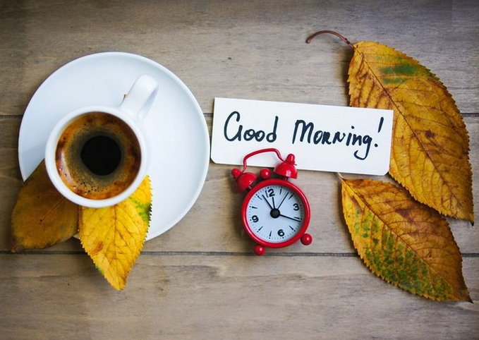 A flat-lay photograph shows a cup of black coffee on a white saucer next to a small red alarm clock and autumn leaves, with a note reading 'Good Morning!' on a wooden surface.