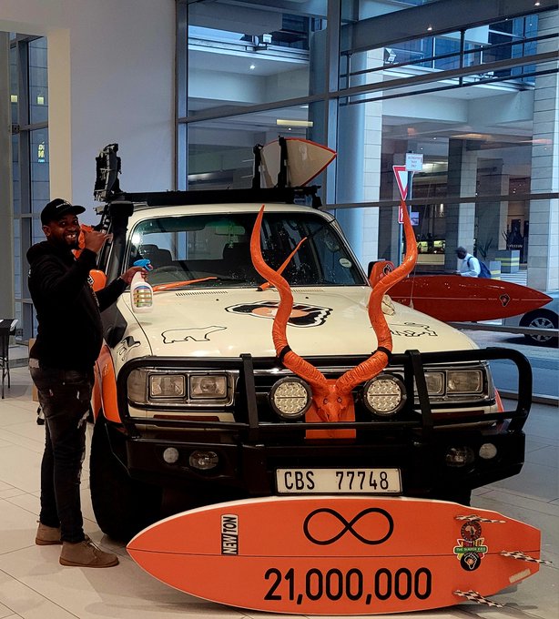 A man in a black cap stands next to a decorated vehicle with orange antlers mounted on the front grille and large orange kudu horns on top. An orange surfboard in front displays 'NEXUSFON', an infinity symbol, a logo, and '21,000,000', with the license plate reading 'CBS 77748'.