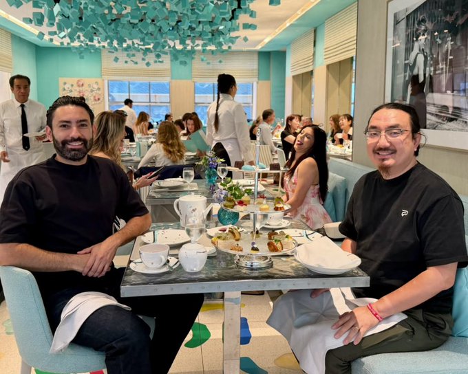 Two men smile while enjoying an elegant afternoon tea with a tiered tray of pastries in a stylish, Tiffany-blue themed restaurant, sharing a warm moment of friendship. The setting—possibly the Tiffany Cafe at The Landmark in London—reflects the celebratory spirit of the caption about gratitude, friendship, and being "Bullish for Meebits."