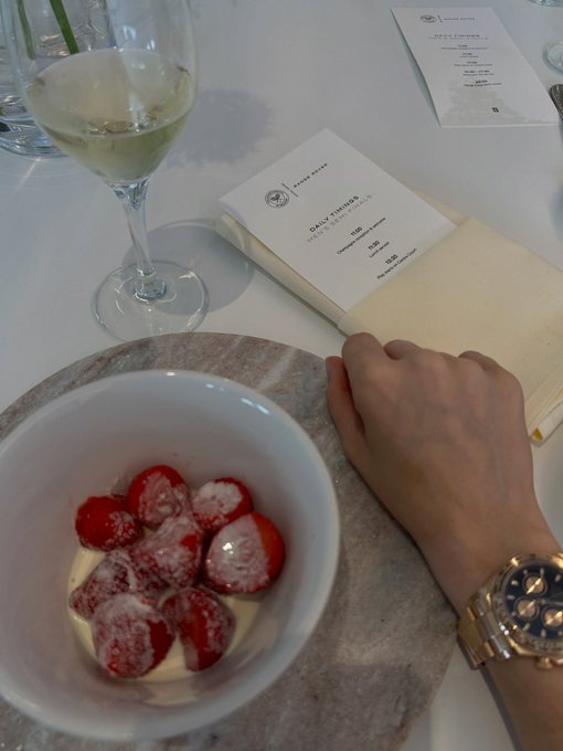 An overhead shot of a restaurant table featuring a bowl of strawberries with cream, a glass of white wine, a person's hand wearing a gold watch, and a receipt marked 'DAILY TIPPING IS NOT A GRATUITY' placed on a notepad.
