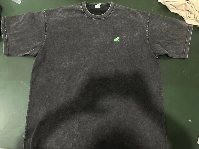 A black acid-wash t-shirt with a small green logo on the chest and a white tag at the neckline lies flat on a dark green surface, appearing to be in vintage or worn condition. The neutral caption "Oh?" doesn't add context to the image, so the relationship is ambiguous—it could be a casual reaction to the retro style or simply a non-sequitur post.