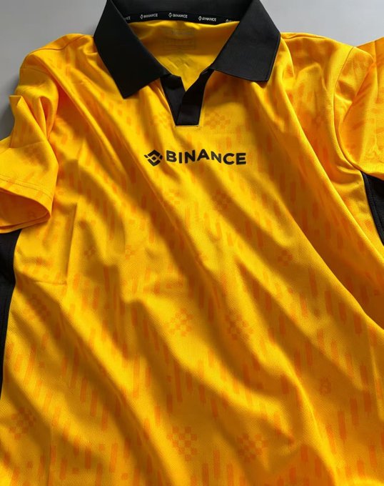 The image features a yellow and black polo shirt with the word "Binance" written across the front. The shirt appears to be new or recently purchased, as indicated by its pristine condition. It's not clear if this particular item of clothing is from Binance or another brand, but it does have a distinctive design that stands out.