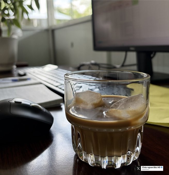 A glass of iced coffee with milk sits on a wooden desk next to a black computer mouse, keyboard, and monitor in a brightly lit workspace.