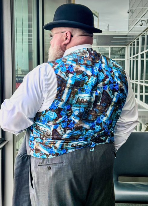 A man with a long blonde beard wearing a bowler hat, white shirt, and a blue floral patterned vest, standing near a window.