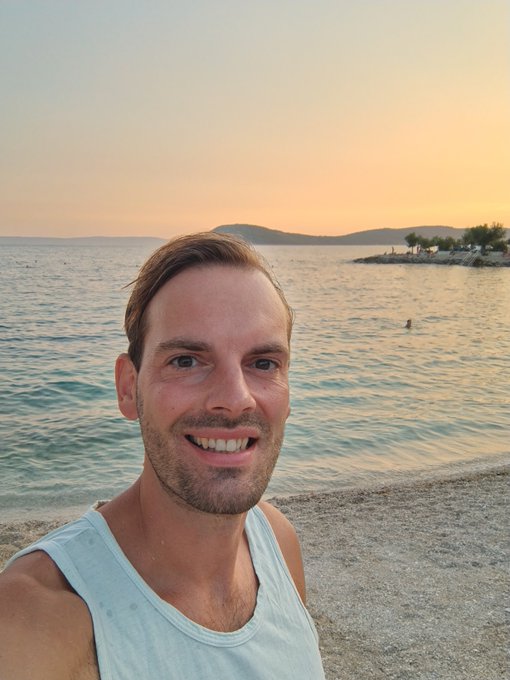 A selfie of a smiling man with short brown hair wearing a light gray tank top, taken on a rocky beach at sunset with calm water and distant mountains in the background.
