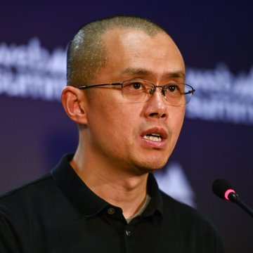 A close-up portrait of Changpeng Zhao (CZ), the former CEO of Binance, speaking at a conference with a microphone in front of him.