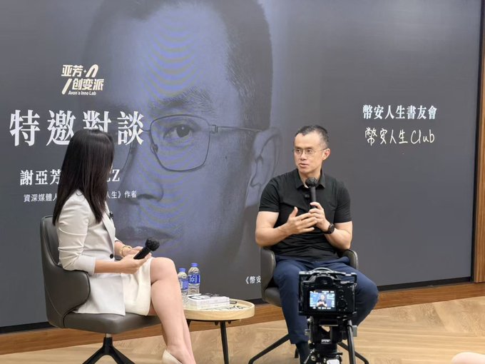 A man and a woman sit on chairs having a discussion on stage, with the woman holding a microphone; a large backdrop behind them features a face and Chinese text including '亞芳の創變派' and '帶安人生書友會'.