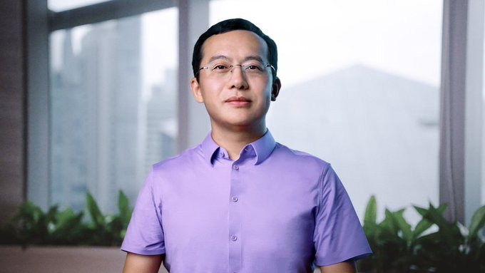 A man wearing a light purple button-up shirt and glasses stands in front of large windows with a cityscape view, likely Star Xu, the founder of OKX, as referenced in the post about the cryptocurrency exchange's European expansion and regulatory milestones.