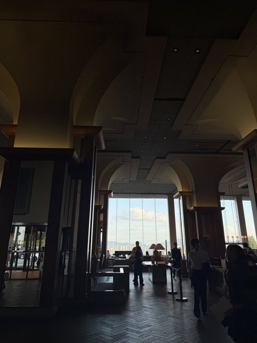 A dimly lit, elegant hotel or building lobby features dark wood columns, arched ceilings, and large windows looking out onto a sunny landscape.