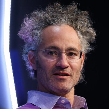 The image shows a man with curly gray hair and glasses, wearing a light-colored collared shirt, appearing to be speaking at an event or conference.