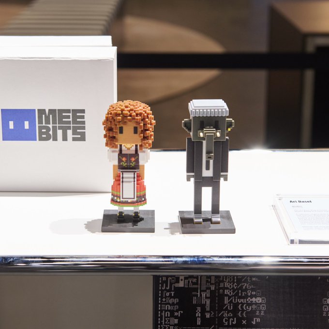 Two blocky, pixel-art style figures displayed on a table next to a 'MEEBITS' sign, with one figure resembling a person with orange hair in a dress and the other a robot-like character.