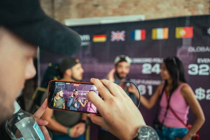 The image shows a man taking a picture of another person with his cell phone at an event. The people present are wearing hats, indicating that they might be attending a special occasion or gathering. There are also several other individuals visible in the background, suggesting that this is a social event where attendees have gathered to interact and enjoy themselves.