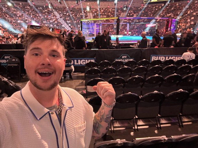 A young man with a beard and sunglasses on his head excitedly poses for a selfie inside a packed UFC arena, with the octagon and "Bud Light" sponsorship banners visible in the background ahead of a fight night event.