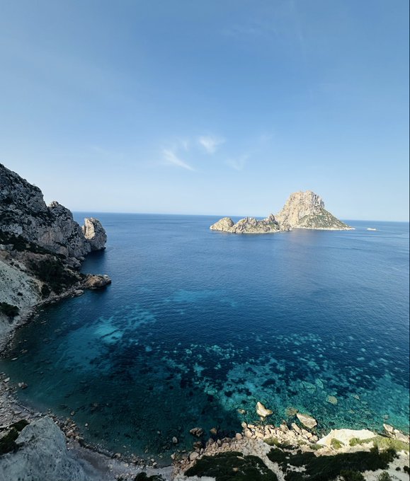 The image shows a beautiful view of an ocean with clear blue water, surrounded by rocky cliffs or mountains. The sky above is also blue, creating a serene atmosphere. In the background, there are some islands visible, adding to the picturesque scenery. The photo appears to be taken from a high vantage point, providing a comprehensive view of the landscape below.