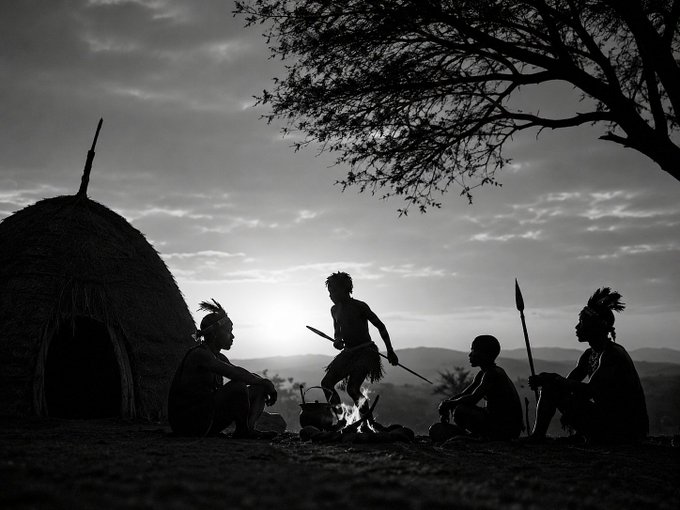 A black and white silhouette photograph showing an indigenous tribe gathered around a campfire near a thatched hut at sunset.