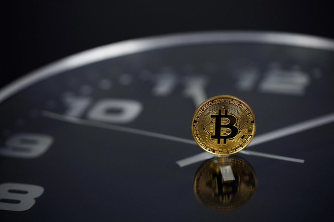 A close-up photograph of a physical gold Bitcoin coin standing upright on a reflective dark surface, with large numbers like '10' and '11' visible in the blurred background.