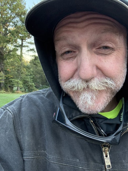 A close-up selfie of an older man with a gray beard and mustache, wearing a dark hooded jacket and a cap, with safety glasses hanging from his collar, set against an outdoor background.
