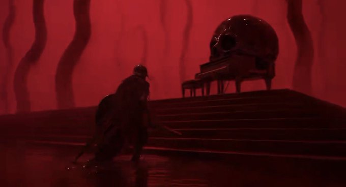 A dark, red-lit scene featuring a silhouetted figure walking up stairs toward a grand piano topped with a large cracked skull.