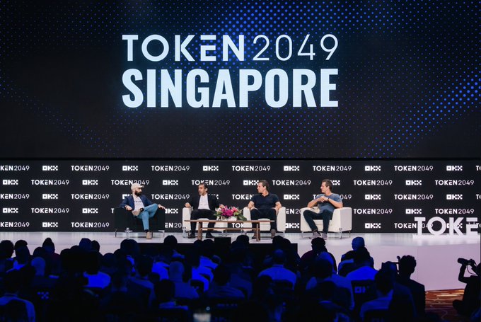 Four panelists sit on stage at TOKEN2049 Singapore, with the event's branding and "OKX" sponsor logos displayed prominently on the backdrop behind them, as they engage in a fireside chat before a large audience.