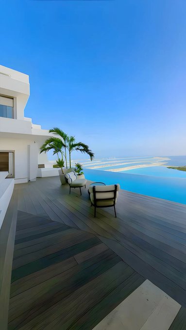A luxurious outdoor terrace with wooden decking, modern lounge chairs, palm trees, and an infinity pool overlooking the ocean with man-made islands visible in the distance.