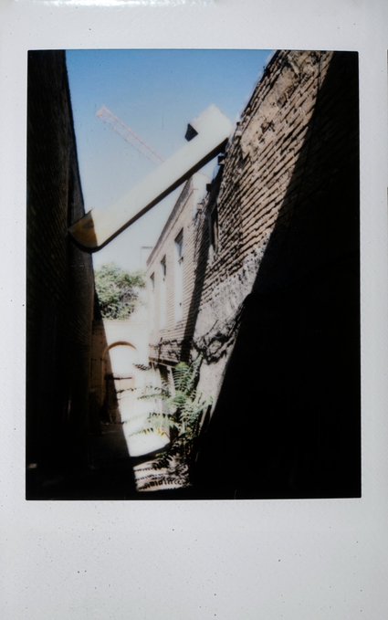 A polaroid-style photograph showing a narrow alleyway between brick buildings, with a long white pipe or beam extending diagonally across the frame and a construction crane visible in the distance against a blue sky.
