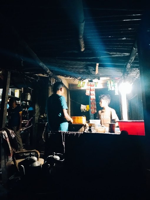 A dimly lit photograph of a small food stall or kitchen at night, with a man in a teal shirt standing at a counter where a shirtless young man is preparing food, illuminated by a single hanging light bulb.