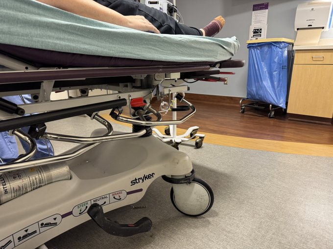 A Stryker-brand hospital gurney with a patient's legs visible on the mattress sits on a vinyl floor in a medical examination room.