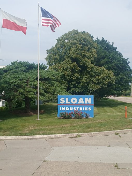 A photograph of a blue sign reading 'SLOAN INDUSTRIES' surrounded by flowers, set on a grassy area with large trees and two flagpoles flying the Polish and American flags.