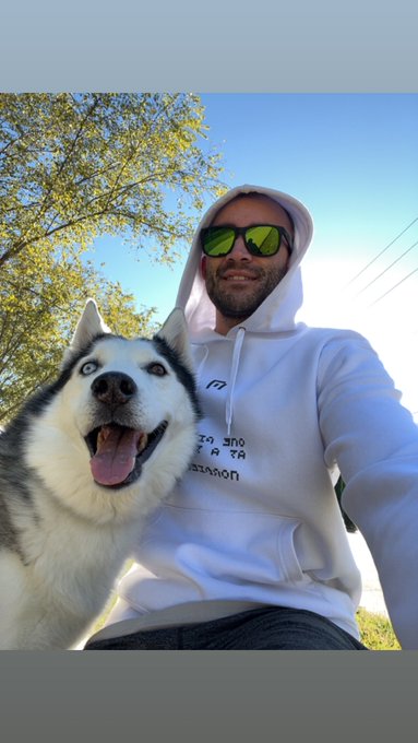 A man wearing sunglasses and a white hoodie poses for a selfie with his happy husky, perfectly capturing "hoodie season" vibes on a sunny fall day.