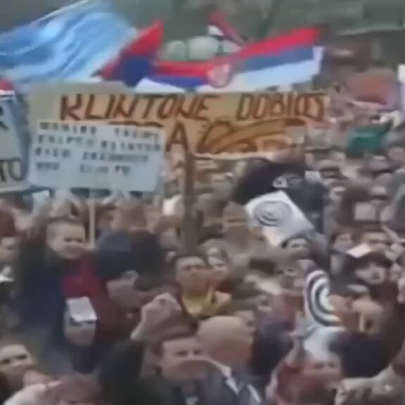 The image shows a large crowd of people gathered together, with some holding signs and flags. The main focus of the scene is a sign that reads "Klintone Dobrios" along with other text. There are also several smaller signs in the background, possibly related to an event or gathering. A person can be seen standing near the center-right part of the image, while another individual is located towards the bottom right corner. The crowd appears to be engaged and interested in what's happening around them.