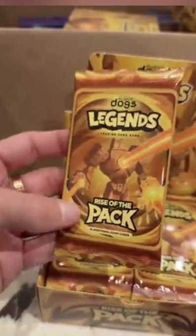 A hand holds a foil pack of trading cards labeled 'All About dogs LEGENDS FEEDING TIME BLEND - RISE OF THE PACK' above a cardboard box filled with similar packs.