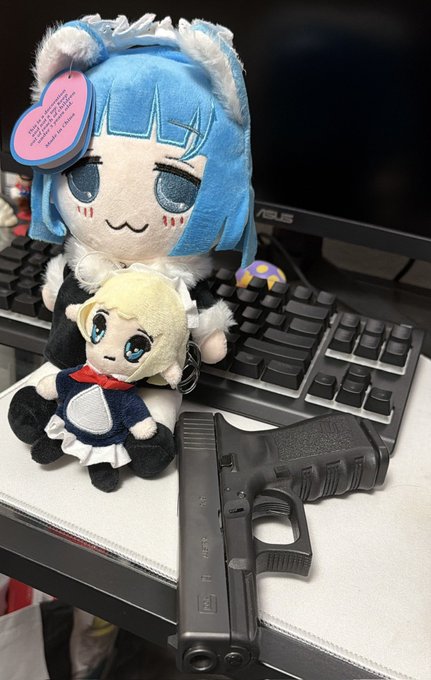 Two anime plush toys sit on a desk next to a black handgun and keyboard; the larger one has blue hair and the smaller one has blonde hair.